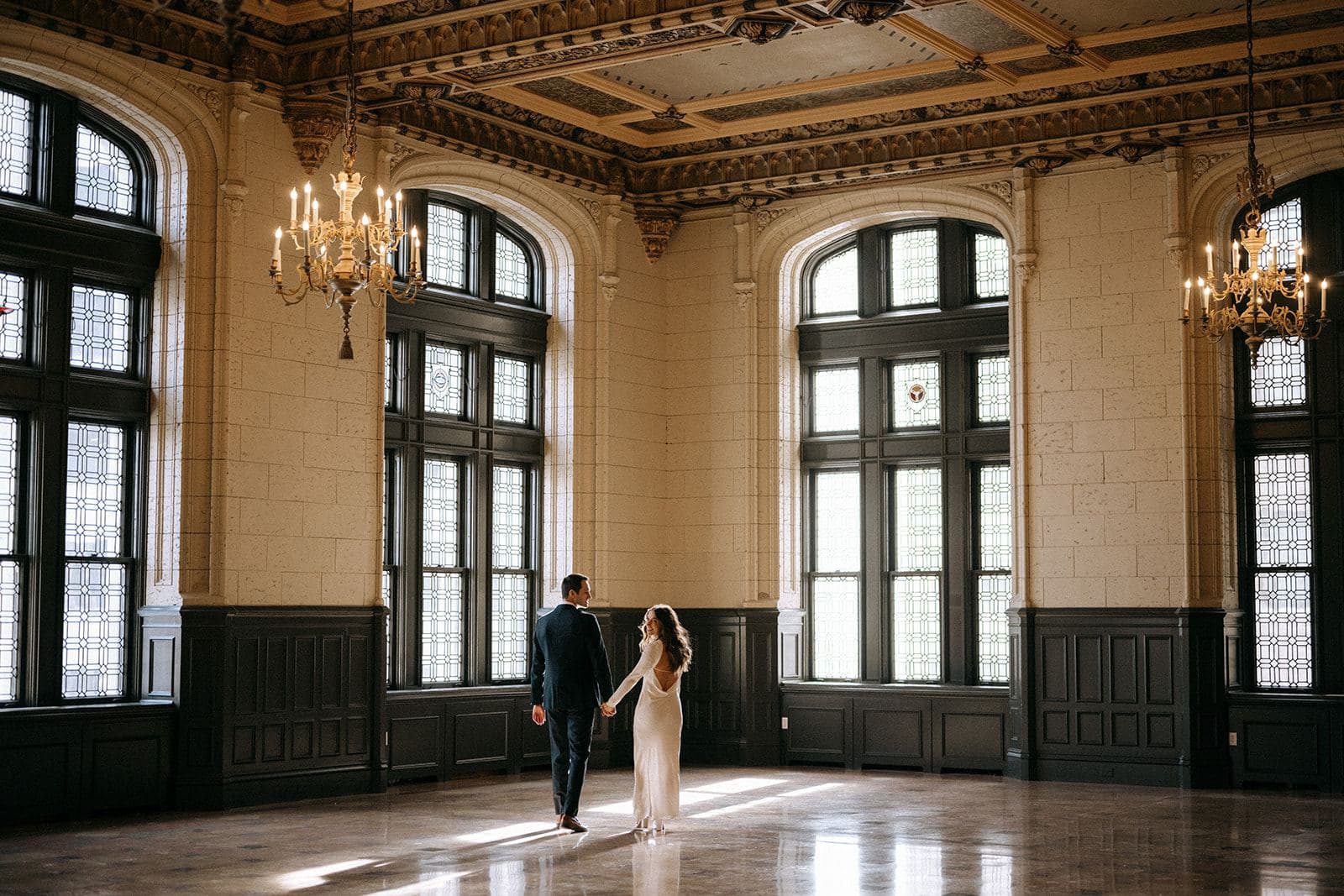 A couple crosses the ballroom floor toward the windows